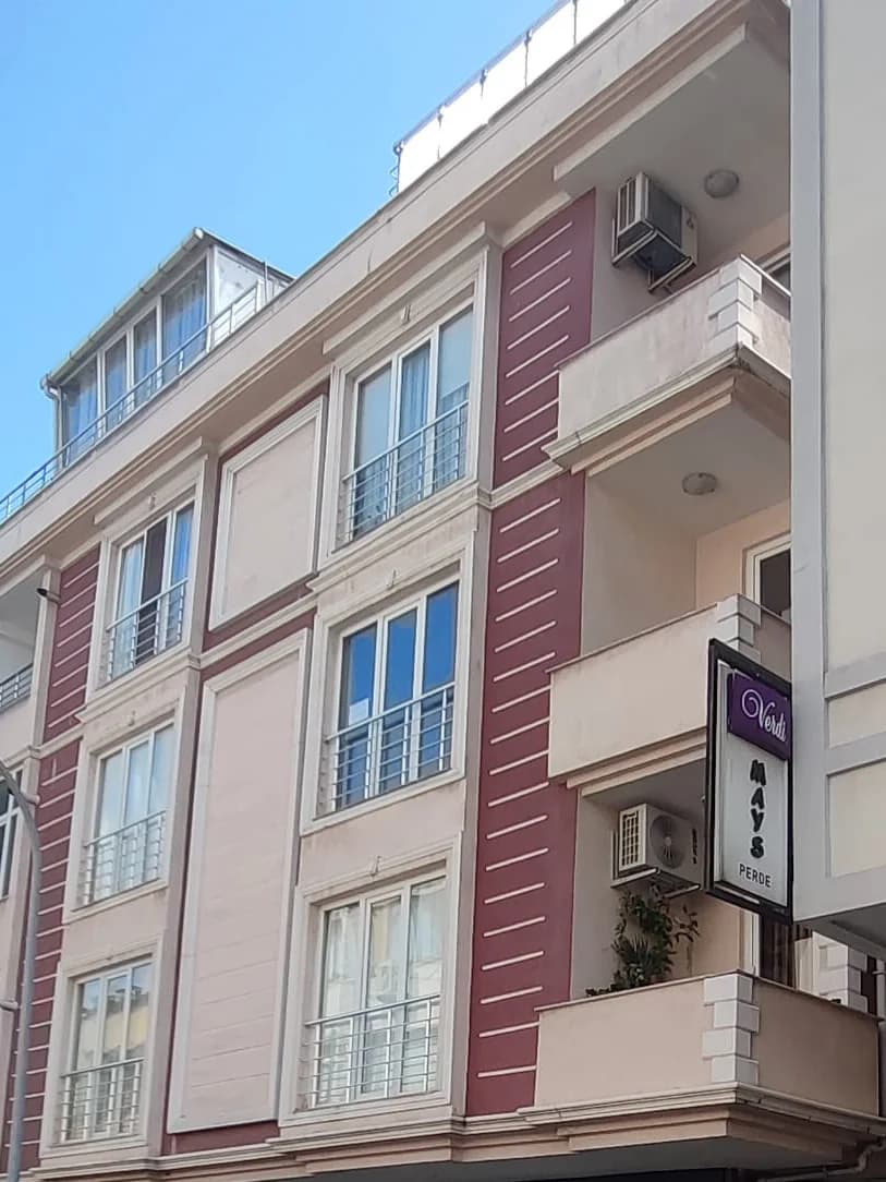 Açık bej cepheli apartmanın üst katları; bordo şeritli yüzeyler, Fransız balkonlu pencereler ve geri çekilmiş camlı çatı katı.