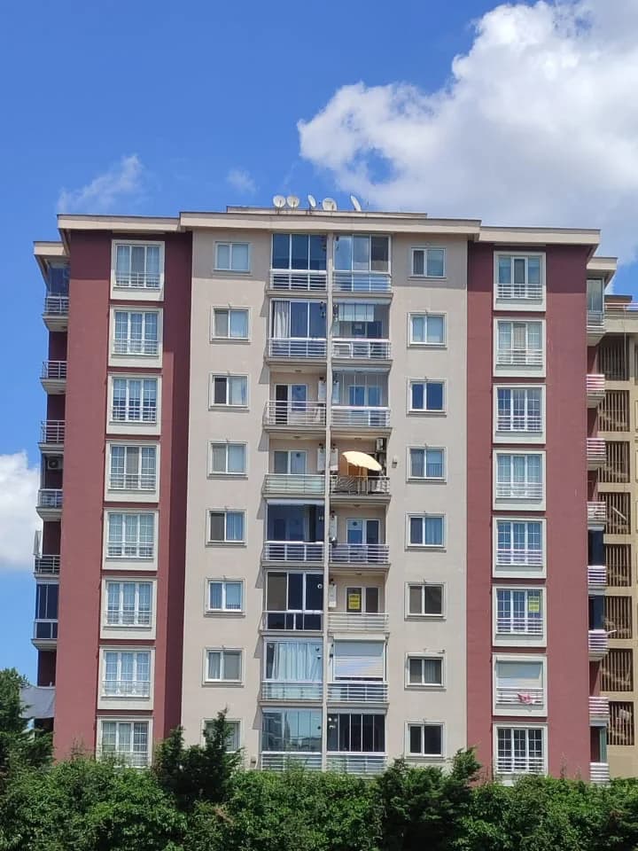 Bordo yan şeritli, açık renkli geniş apartman cephesi; orta aksta camla kapatılmış balkonlar.
