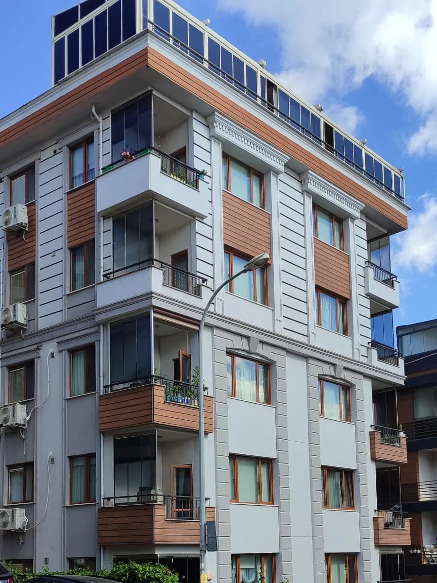 Gri cepheli, ahşap görünümlü kaplama şeritli köşe apartmanı; en üstte camla çevrili çatı katı.