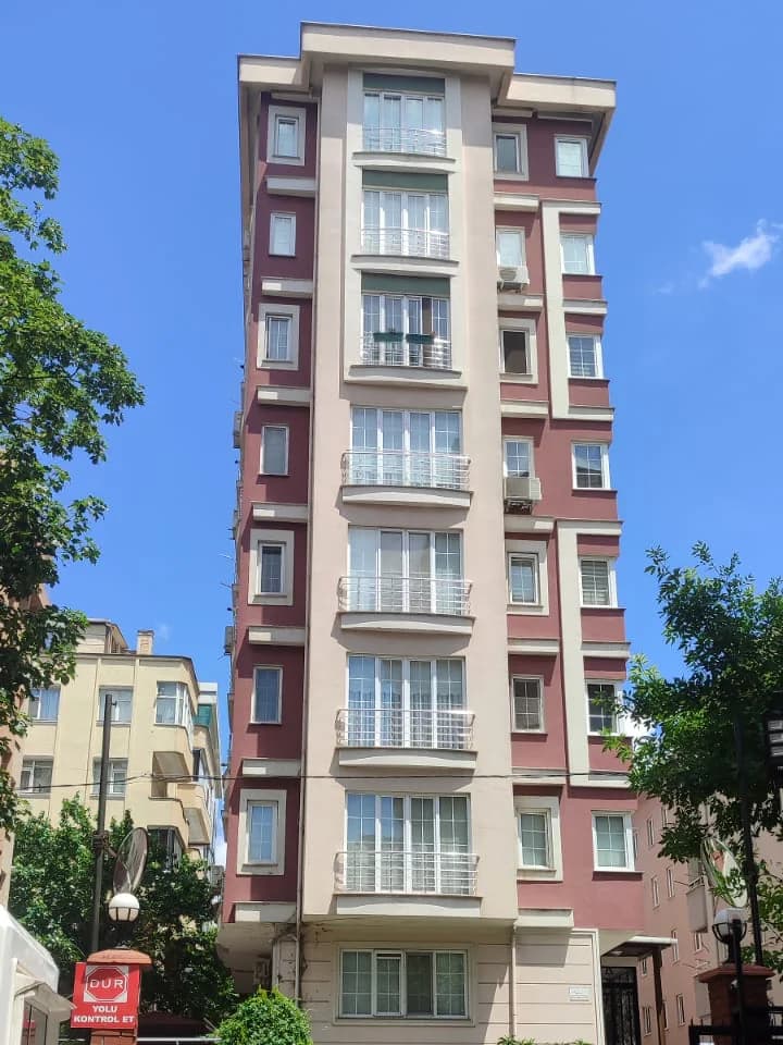 Bordo ve bej dikey şeritli, dar ve yüksek apartman cephesi; ortada üst üste sıralanan geniş pencereler ve beyaz korkuluklar, iki yanda ağaçlar.