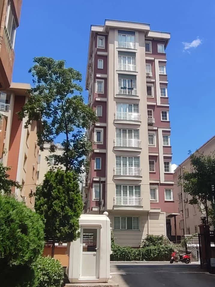 Ağaçların arasından görünen apartman; bordo-bej cephe boyunca üst üste sıralanan balkonlar.
