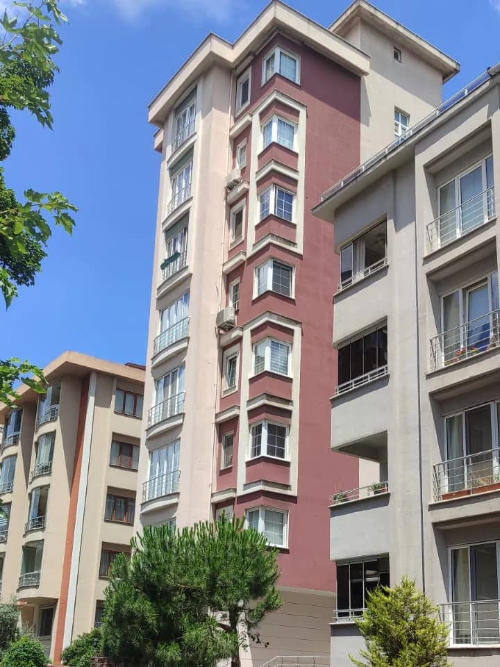 Apartmanın üst katları ve sağda daha alçak komşu yapı, bulutlu gökyüzü önünde.