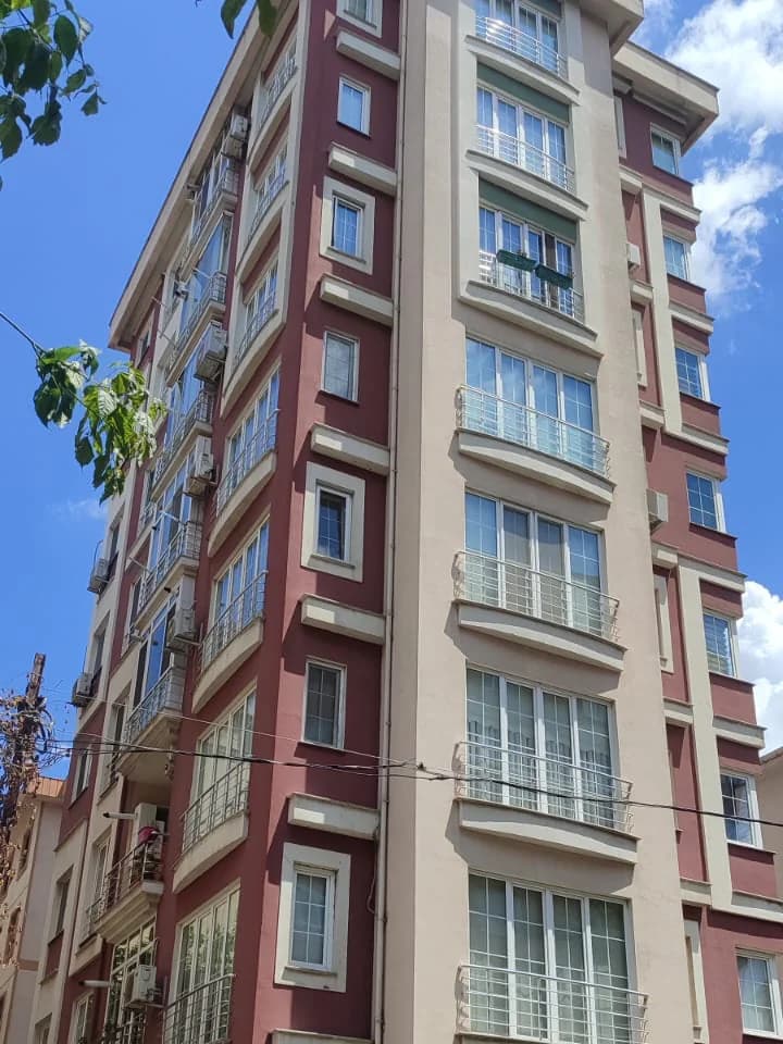Sağ köşeden aşağıdan bakış: geniş pencerelerin ve beyaz korkulukların sıralandığı bordo-bej cephe, sağda bulutlar.