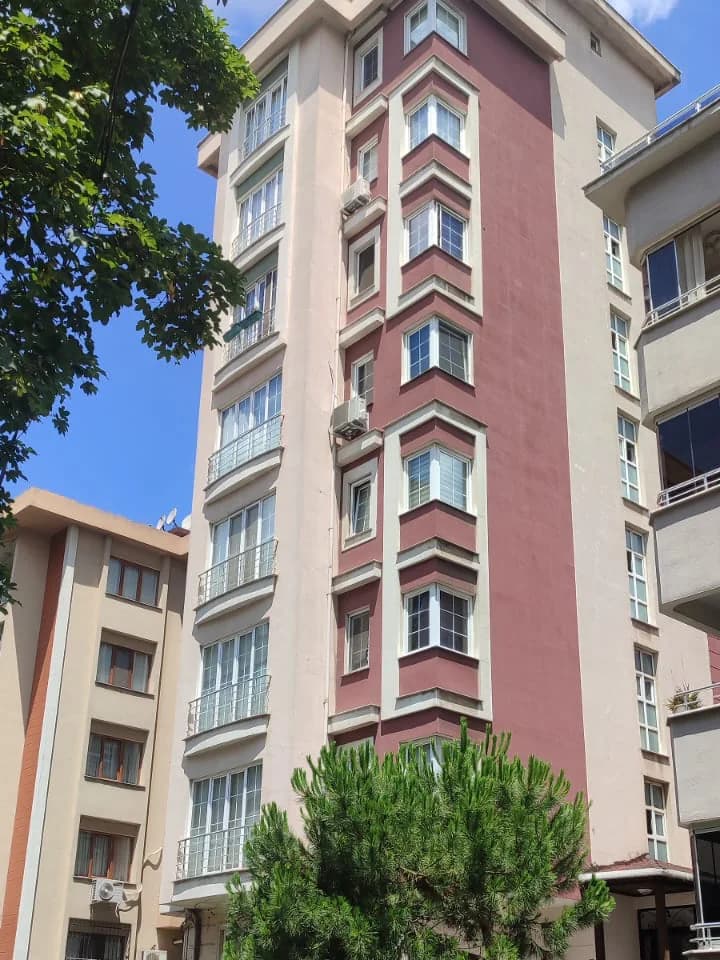 Köşe çıkmaları ve pencere dizisi; solda alçak komşu yapı, altta ağaç.